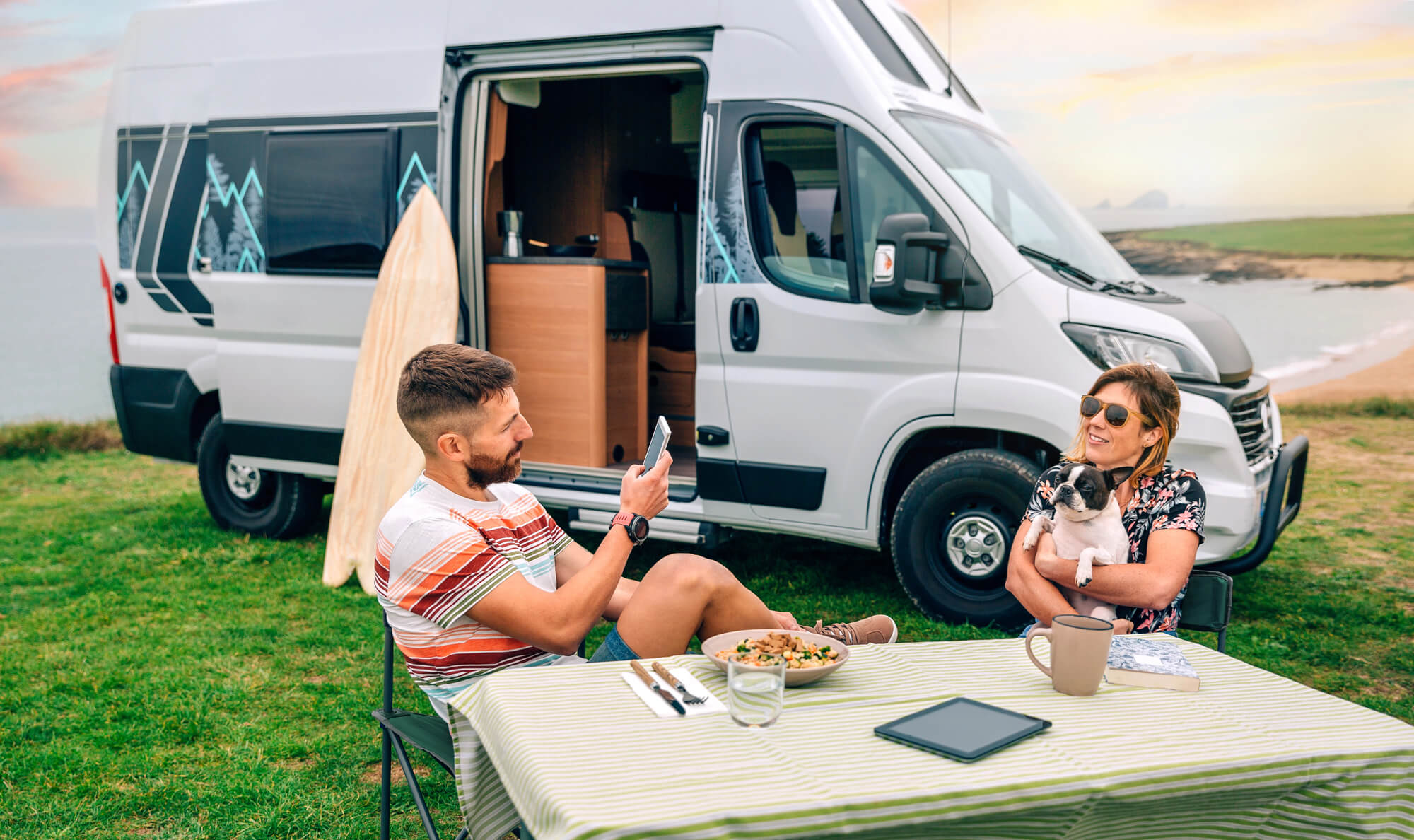 Close-up of a couple with their dog having coffee, with an LWB campervan in the background.